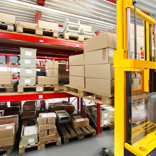 In einer Lagerhalle stapeln sich Kartons und Pakete auf Holzpaletten und roten Metallregalen. Rechts im Vordergrund ist eine gelbe Hebemaschine zu sehen.