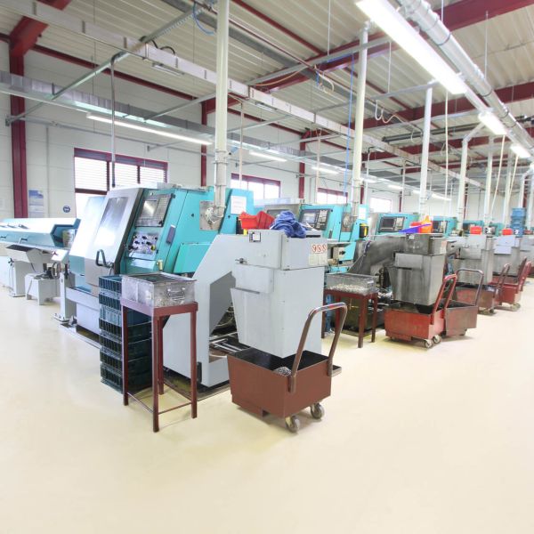 Ein Arbeiter im roten Overall bedient eine große, moderne CNC-Maschine in einer hellen Industriewerkstatt. Im Hintergrund sind ähnliche Maschinen und ein weiterer Arbeiter zu sehen.