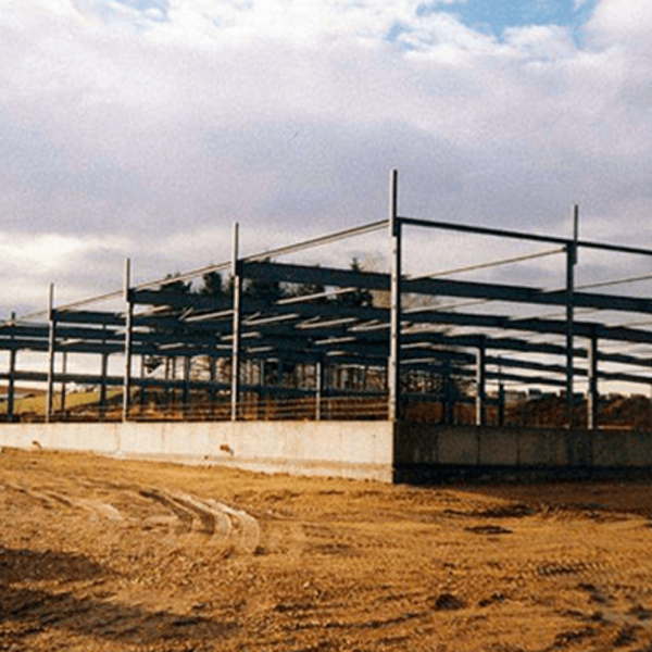Ein Stahlgerüst eines im Bau befindlichen Gebäudes steht auf einem unbefestigten Grundstück unter einem teilweise bewölkten Himmel, wobei weder Wände noch Dach fertiggestellt sind.