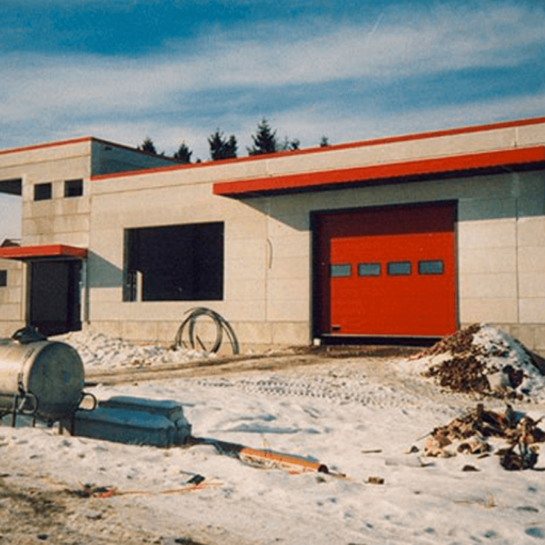 Ein modernes Industriegebäude mit beigen Wänden und einem großen roten Garagentor, umgeben von Schnee und Baumaterialien unter einem teilweise bewölkten Himmel.