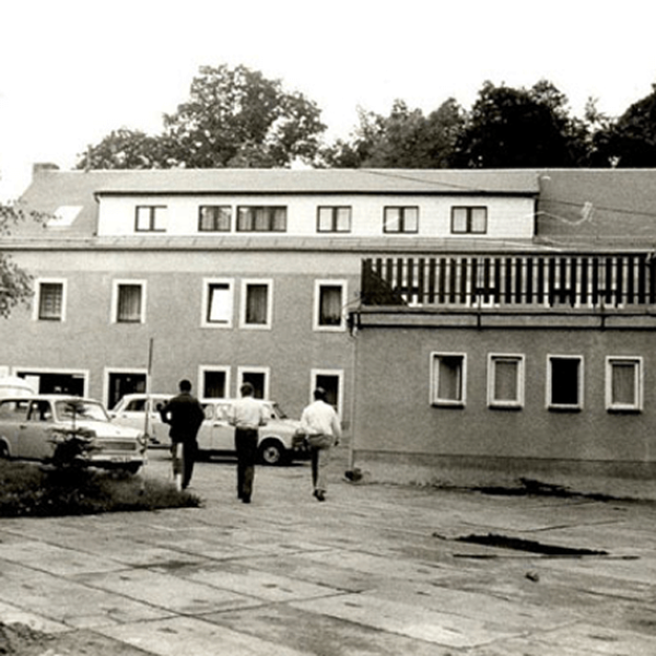 Drei Personen gehen auf ein zweistöckiges Gebäude mit Flachdach und mehreren Fenstern zu; in der Nähe sind einige Oldtimer geparkt. Das Bild ist in schwarz-weiß gehalten. Im Hintergrund sind Bäume zu sehen.