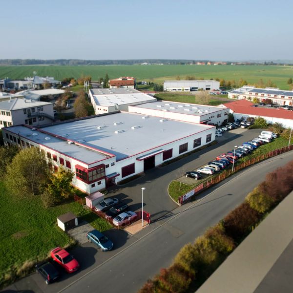 Luftaufnahme eines großen Industrie- oder Bürogebäudes mit weißem Dach, umgeben von geparkten Autos, Grünflächen und anderen Gebäuden in der Nähe in einer ländlichen Gegend.