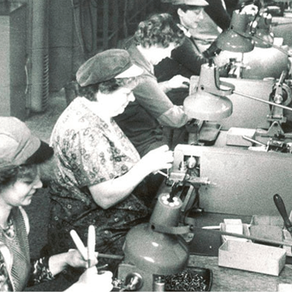 Ein Schwarz-Weiß-Foto zeigt mehrere Frauen, die an Werkbänken sitzen, Maschinen bedienen und unter Schreibtischlampen in einer Fabrik kleine Teile zusammenbauen.