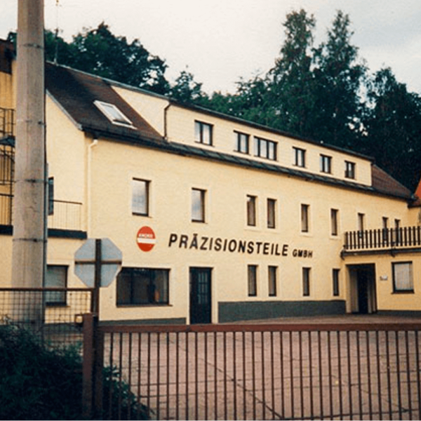 Ein gelbes Gebäude mit der Aufschrift PRÄZISIONSTEILE GMBH an der Fassade, umgeben von Bäumen und einem Zaun, mit einem Stoppschild und einer Ecke eines Tores im Vordergrund.
