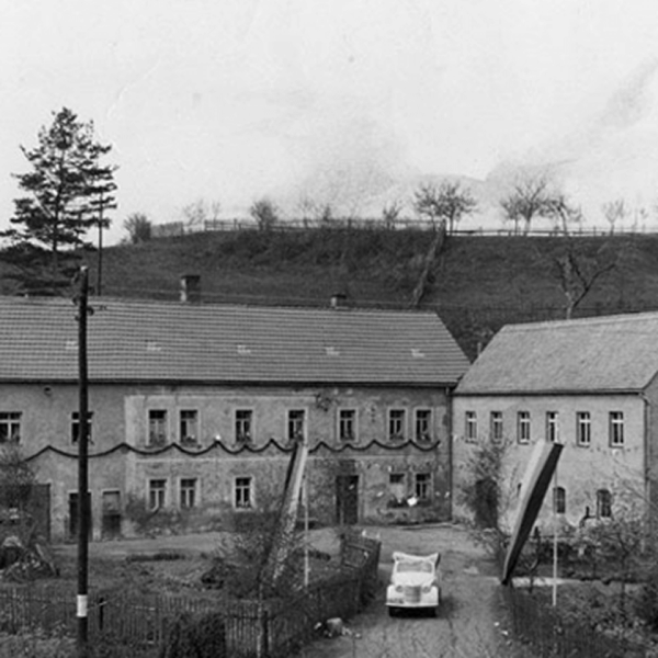 Schwarz-Weiß-Foto einer ländlichen europäischen Szene mit zwei alten Gebäuden, einem Zaun, kahlen Bäumen und einem kleinen Auto, das in der Mitte eines Hofes geparkt ist. Im Hintergrund ist ein grasbewachsener Hügel zu sehen.