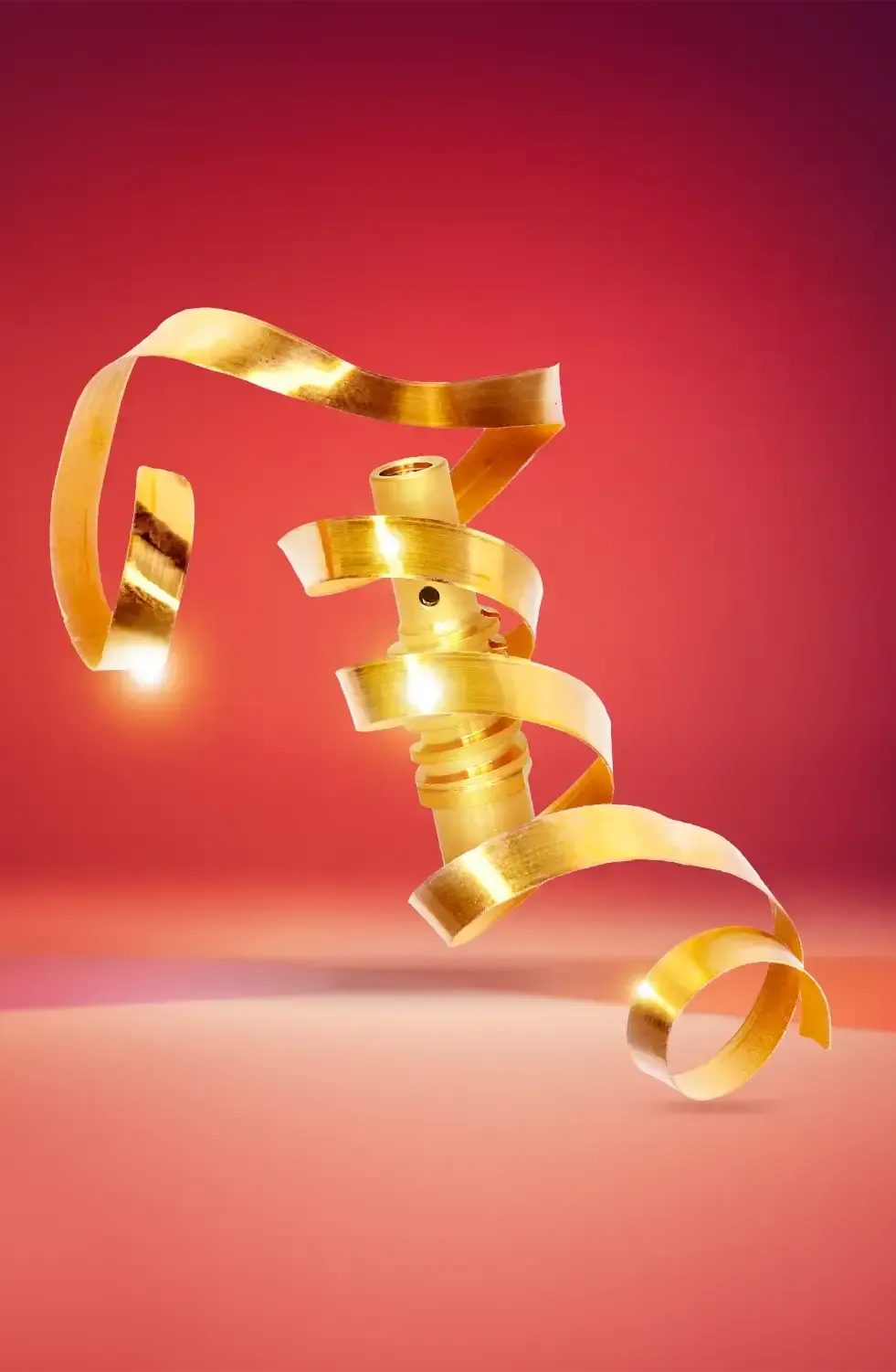 Ein goldglänzendes Band windet sich spiralförmig um ein kleines, zylindrisches Metallobjekt, das vor einem rosa und roten Hintergrund mit Farbverlauf leuchtet. Die Szene ist hell beleuchtet und hebt die glatte, reflektierende Textur des Bandes hervor.