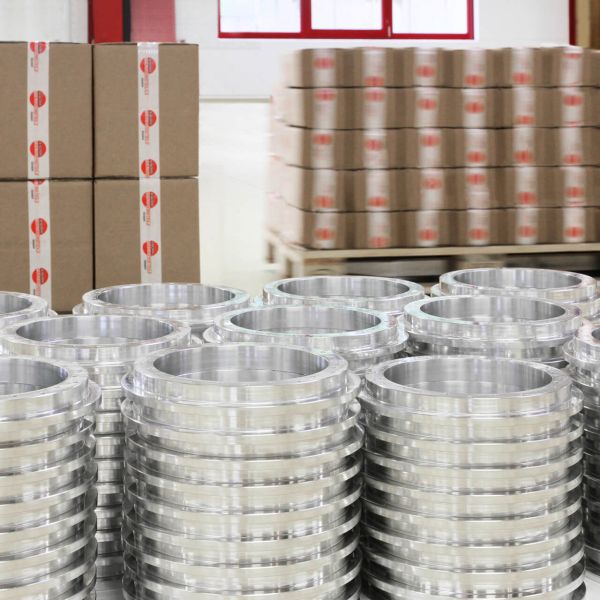 In einer Lagerhalle stapeln sich Kartons und Pakete auf Holzpaletten und roten Metallregalen. Rechts im Vordergrund ist eine gelbe Hebemaschine zu sehen.