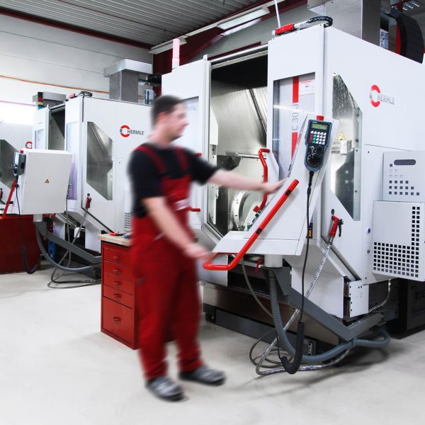 Ein Arbeiter im roten Overall bedient eine große, moderne CNC-Maschine in einer hellen Industriewerkstatt. Im Hintergrund sind ähnliche Maschinen und ein weiterer Arbeiter zu sehen.